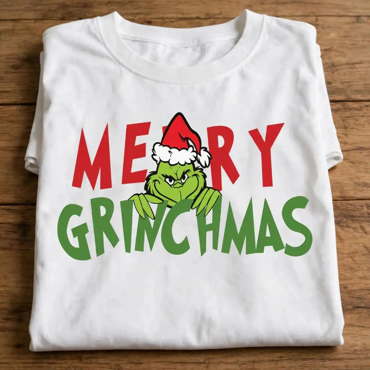 merry grinchmas png design with the grinch and santa hat a green furry creature wearing a Santa hat, peeking over green text, with red text above design printed on a plain product mo